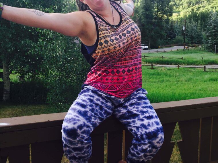 A person with outstretched arms and open mouth sits barefoot on a wooden railing, wearing a colorful tank top and tie-dye pants; green hills and trees are in the background—celebrating like they’ve found indoor plumbing for the gold!. Soul Flower Blog: Hippie Style & Kind Living