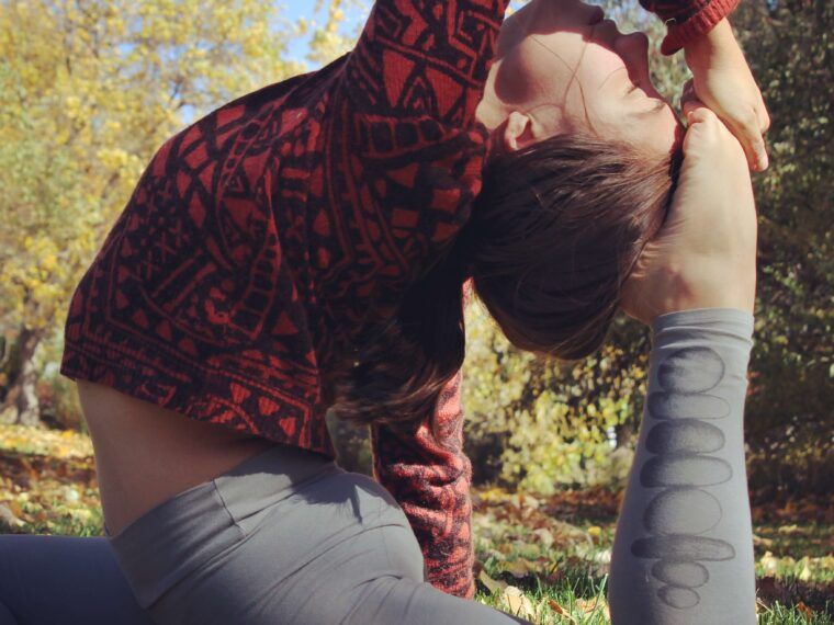 Person practicing a deep yoga stretch outdoors on grass, wearing a funky red sweater and gray leggings, with trees and blue sky in the background for an eco-inspired vibe. Soul Flower Blog: Hippie Style & Kind Living