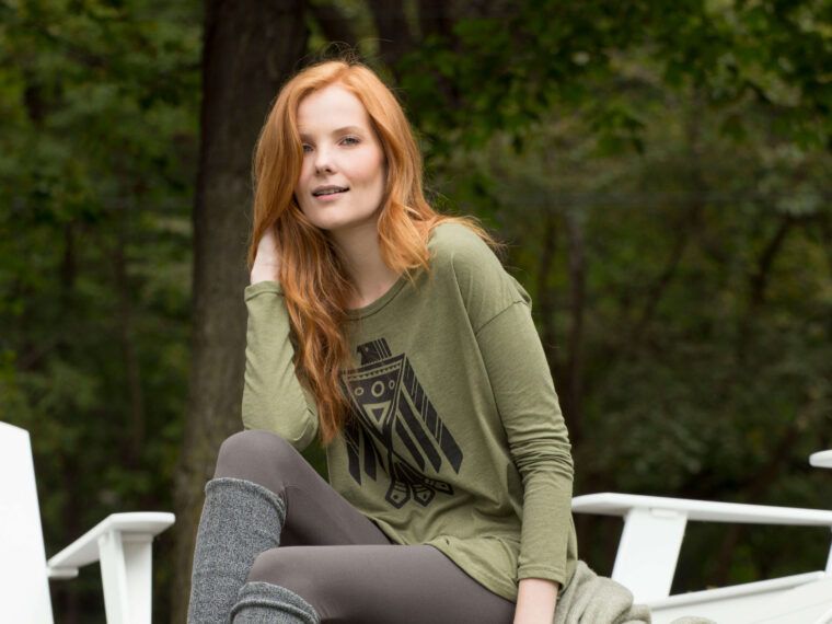 A woman with long red hair sits outdoors on a white chair, wearing an earthy green hippie t-shirt featuring a black geometric bird design, gray leggings, and leg warmers. Trees and greenery form the tranquil backdrop. Soul Flower Blog: Hippie Style & Kind Living