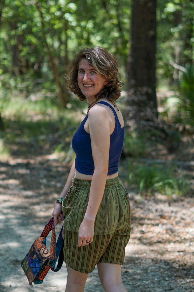 Model wearing navy blue crop tank with striped green hippie shorts standing in forest smiling.