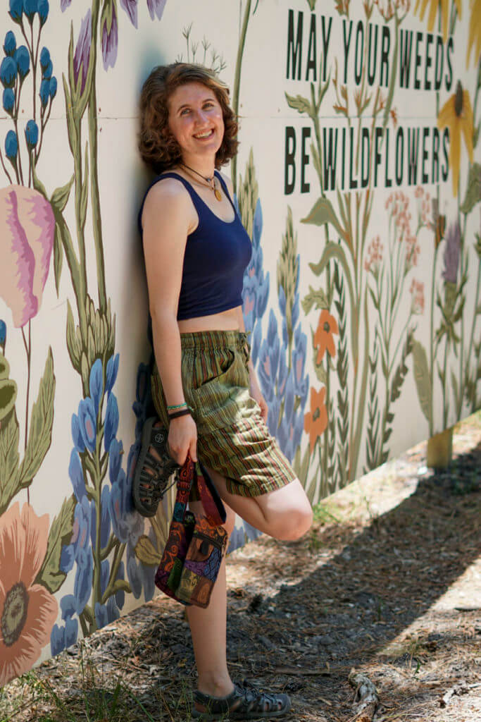 Model wearing navy blue crop tank with striped green hippie shorts standing in forest smiling.