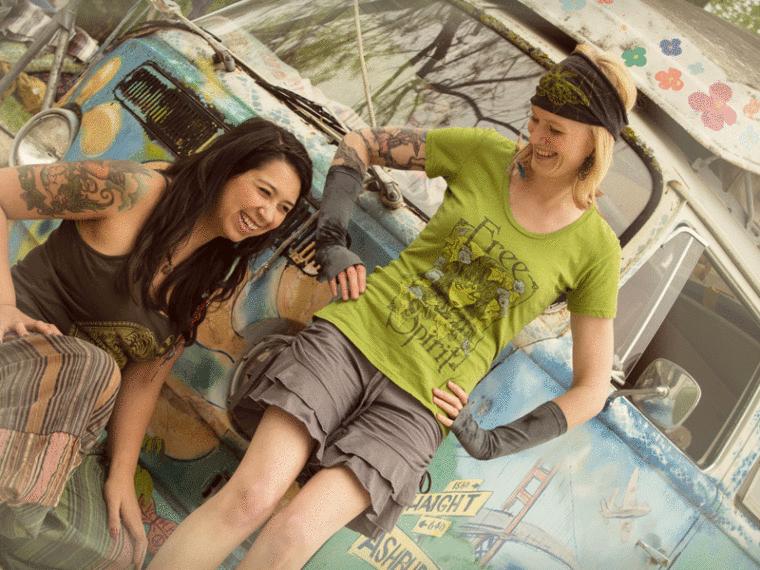 Two women with tattoos smile and lean against a colorful, painted van outdoors. One wears a green Free Spirit shirt; both appear relaxed and happy, radiating an eco-boho vibe perfect for any free-spirited, hippie adventure. Soul Flower Blog: Hippie Style & Kind Living