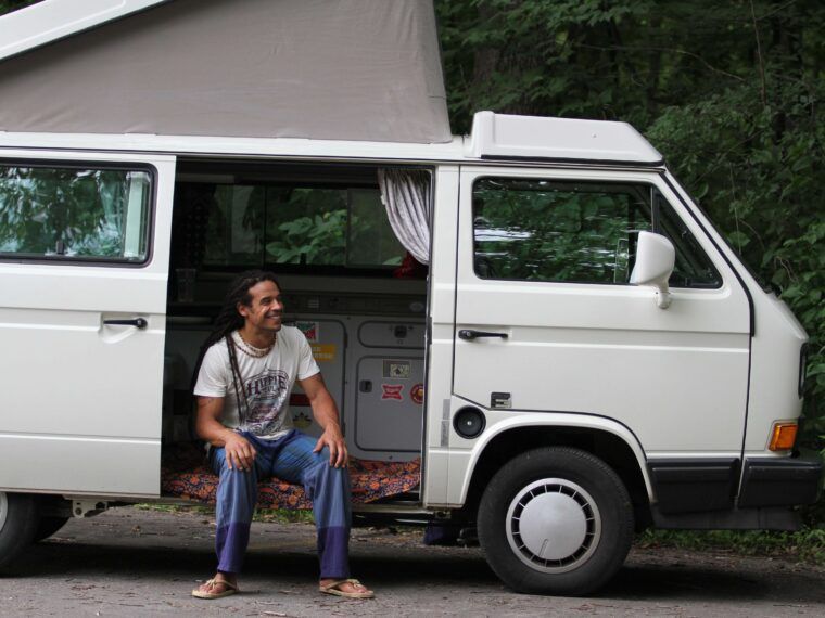 A person sits in the doorway of a white camper van parked on a road, surrounded by trees and greenery—an inspiring setting for reflecting on 10 Ways to Celebrate the Fall Equinox. Soul Flower Blog: Hippie Style & Kind Living