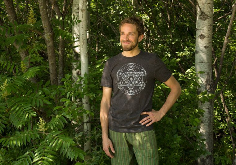 A man stands in a forested area with one hand on his hip, wearing a dark geometric-print t-shirt and green striped pants—ready to kickstart his week with a Monday Motivation Playlist. Soul Flower Blog: Hippie Style & Kind Living