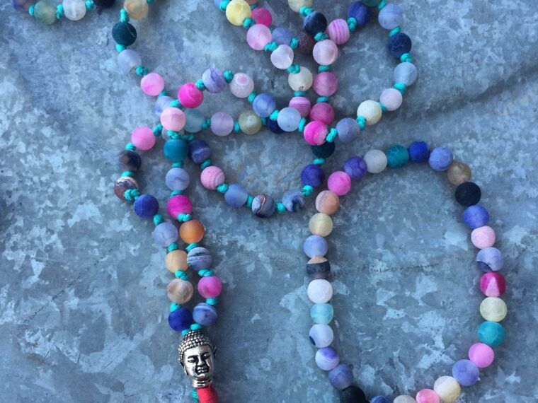 A beaded necklace from Mantra & Malas with multicolored round beads, a silver Buddha charm and red tassel, and another featuring a silver Om charm with blue tassel, elegantly displayed on a stone surface. Soul Flower Blog: Hippie Style & Kind Living