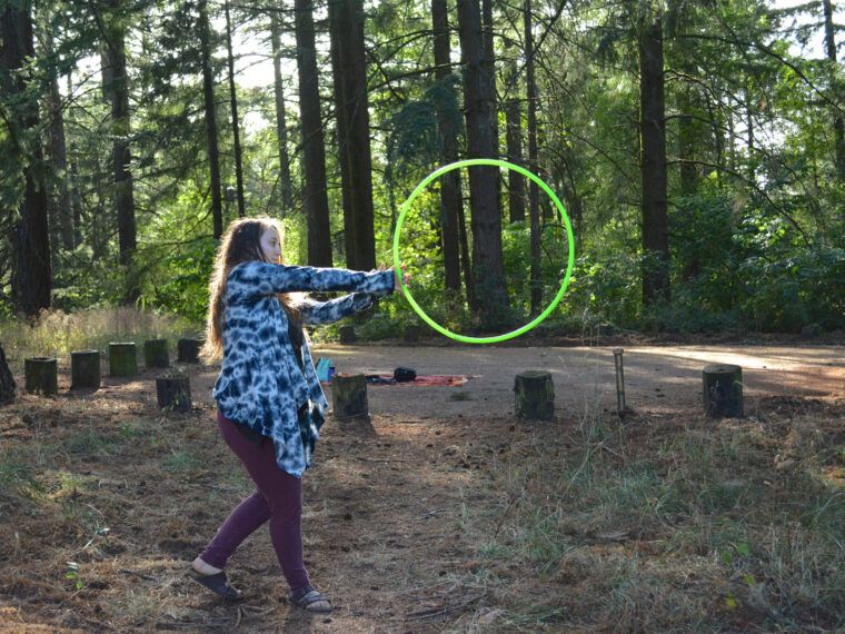 A person with a funky vibe stands in a forested area, holding a large green hoop on dry ground, surrounded by an earthy circle of wooden stumps. Soul Flower Blog: Hippie Style & Kind Living