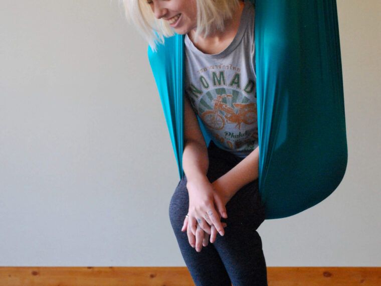 A person with blonde hair sits in an earthy teal aerial yoga hammock, smiling, with legs crossed, on a blue mat indoors. The text SOUL FLOWER appears in the bottom right corner. Soul Flower Blog: Hippie Style & Kind Living