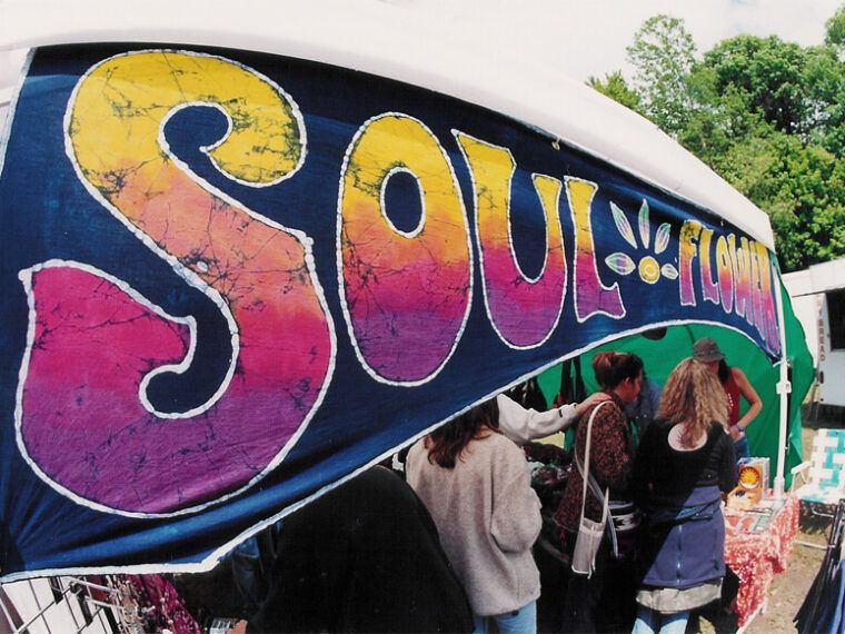 A market stall with a colorful tie-dye banner reading SOUL FLOWER welcomes shoppers for Small Business Saturday, with several people browsing unique items on tables under the tent. Soul Flower Blog: Hippie Style & Kind Living