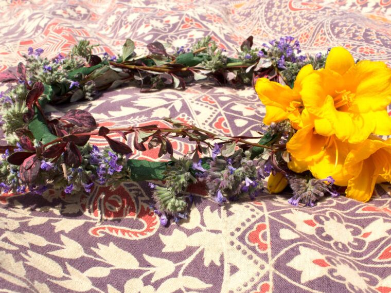 A Hippie Flower Crown made of yellow flowers, purple blossoms, and greenery lies on a patterned fabric background. Soul Flower Blog: Hippie Style & Kind Living