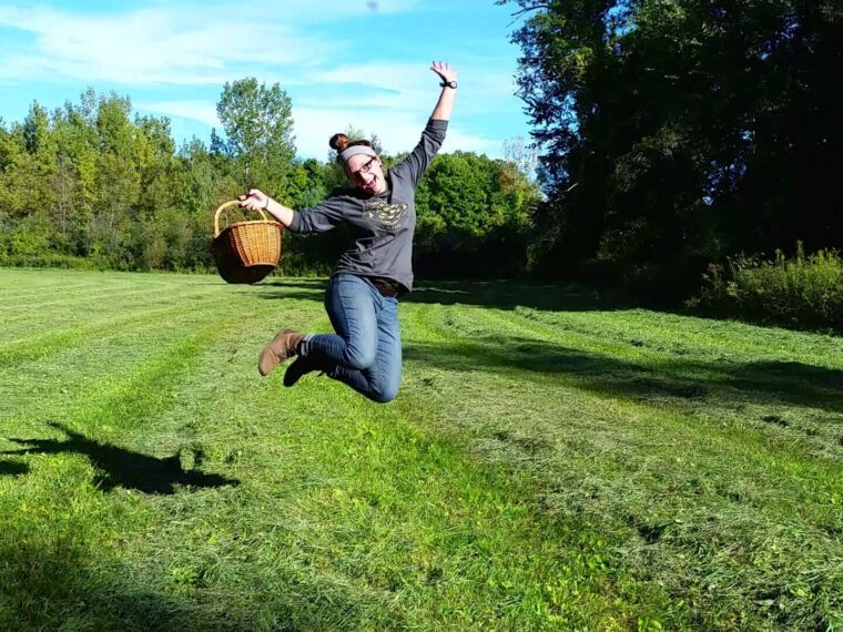 A person with a bun hairstyle jumps in the air on a grassy field, holding a wicker basket; trees and blue sky set the scene, echoing the spirit of "You Belong Among the Wild Fox Grapes. Soul Flower Blog: Hippie Style & Kind Living