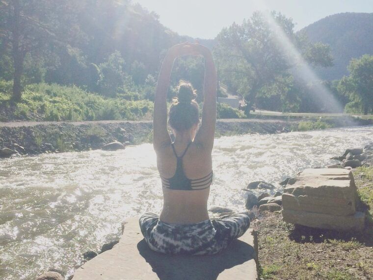 Person sitting cross-legged on a rock by a river, stretching arms overhead, with sunlight streaming through trees—capturing the calm you’ll feel by following these 7 Tips for Having a Stress-Free Wedding Day. Soul Flower Blog: Hippie Style & Kind Living