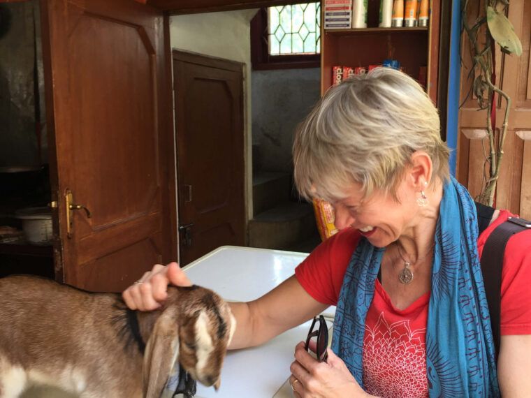A woman with a boho vibe smiles while petting a young goat indoors, next to a white appliance, with shelves and an orange wall in the background. Soul Flower Blog: Hippie Style & Kind Living