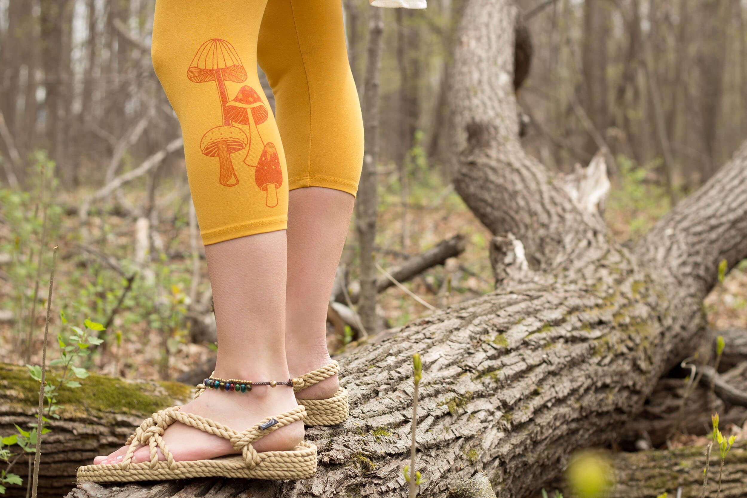 A person wearing yellow leggings with orange mushroom prints and rope sandals stands on a fallen tree in a forest—maybe a nod to the question, Why are Mushrooms Everywhere Right Now? Only their lower legs and feet are visible.