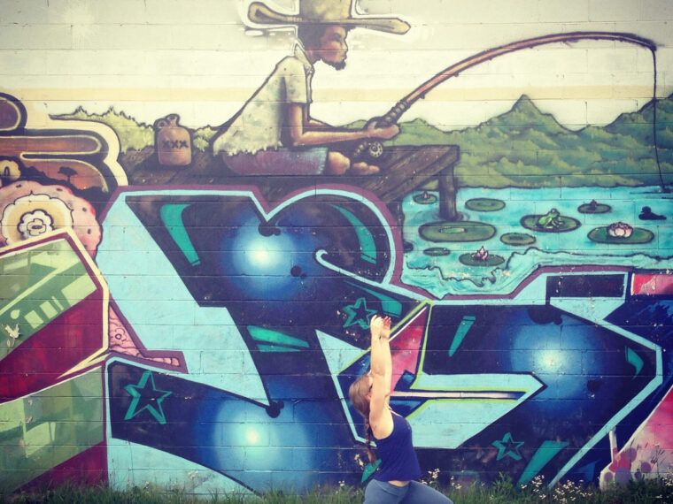 A person does a yoga pose in front of a colorful, earthy mural depicting a person fishing by a lake, with boho graffiti and landscape elements. Soul Flower Blog: Hippie Style & Kind Living