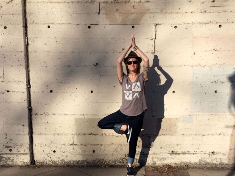 A person wearing sunglasses and a t-shirt stands in a yoga tree pose against a concrete wall, radiating an earthy, hippie vibe, with their shadow visible behind them. Soul Flower Blog: Hippie Style & Kind Living