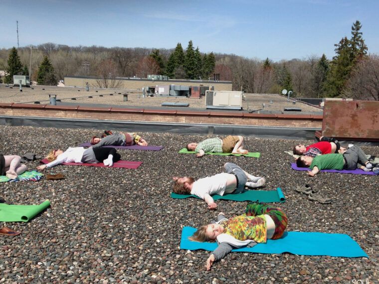 A group of people lying on yoga mats, practicing relaxation or meditation on an earthy rooftop covered with small stones, under a clear sky. Soul Flower Blog: Hippie Style & Kind Living