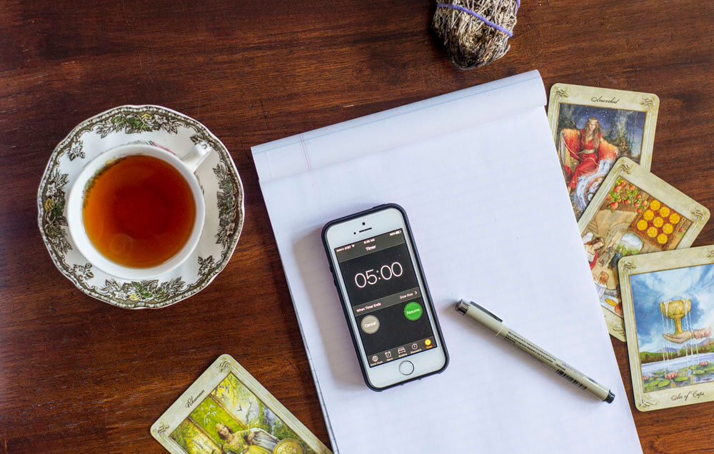 A cup of tea, a pen, a smartphone displaying a five-minute timer, tarot cards, and a notepad rest on a wooden table—perfect for mindful living with a hint of boho charm. Soul Flower Blog: Hippie Style & Kind Living