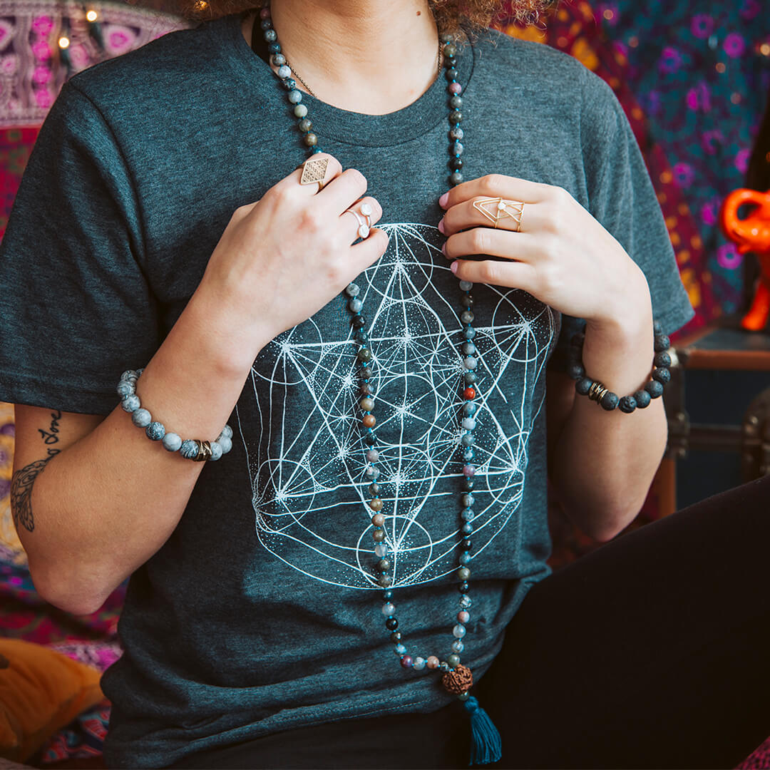 Person wearing a dark geometric print t-shirt featuring the Merkaba Symbol, beaded necklaces, rings, and bracelets, with colorful patterned fabric in the background. Soul Flower Blog: Hippie Style & Kind Living