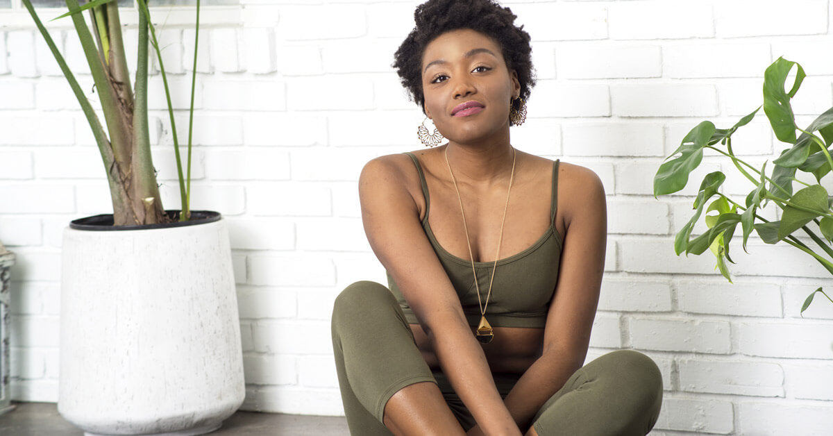 Woman in olive green workout clothes sits on the floor against a white brick wall, surrounded by potted plants—embracing mindful living and a natural, eco-inspired space. Soul Flower Blog: Hippie Style & Kind Living