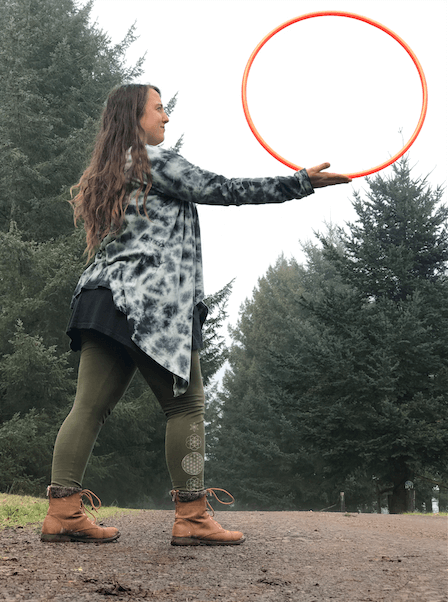 A person with a boho vibe stands outdoors on a path, holding a large orange hoop upright with one hand, surrounded by tall evergreen trees. Soul Flower Blog: Hippie Style & Kind Living