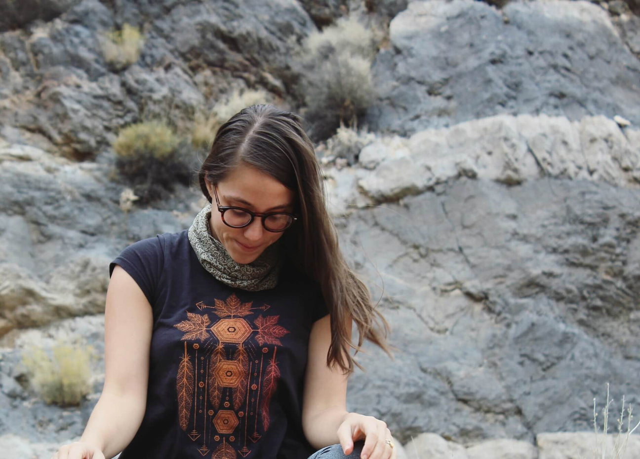 A person with long hair, glasses, and a patterned shirt sits outdoors in front of rocky terrain, embracing a boho vibe as they look down thoughtfully. Soul Flower Blog: Hippie Style & Kind Living