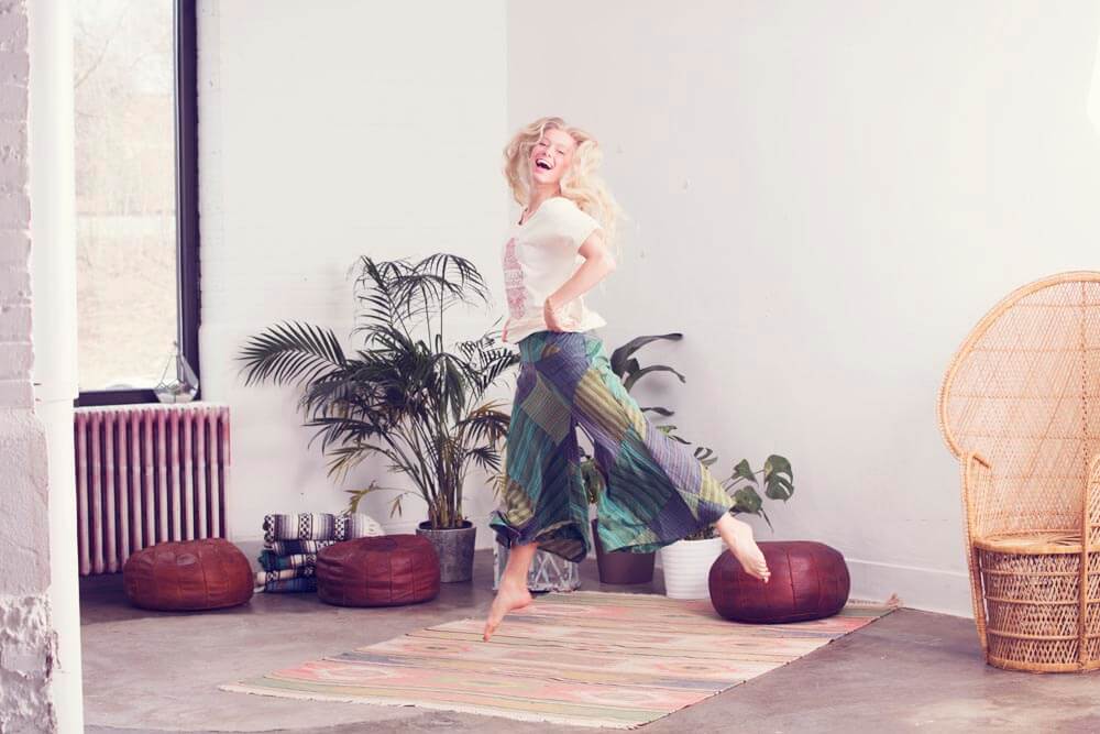 A woman with long blonde hair jumps joyfully in a bright, natural room with boho plants, poufs, and a woven chair, standing on a striped rug near a large window. Soul Flower Blog: Hippie Style & Kind Living