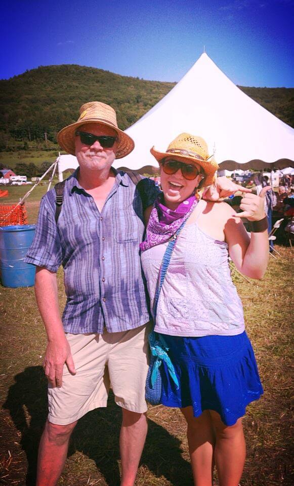 Two people in boho summer clothing and hats stand outdoors at a festival, with a large white tent and green hills in the background. Soul Flower Blog: Hippie Style & Kind Living