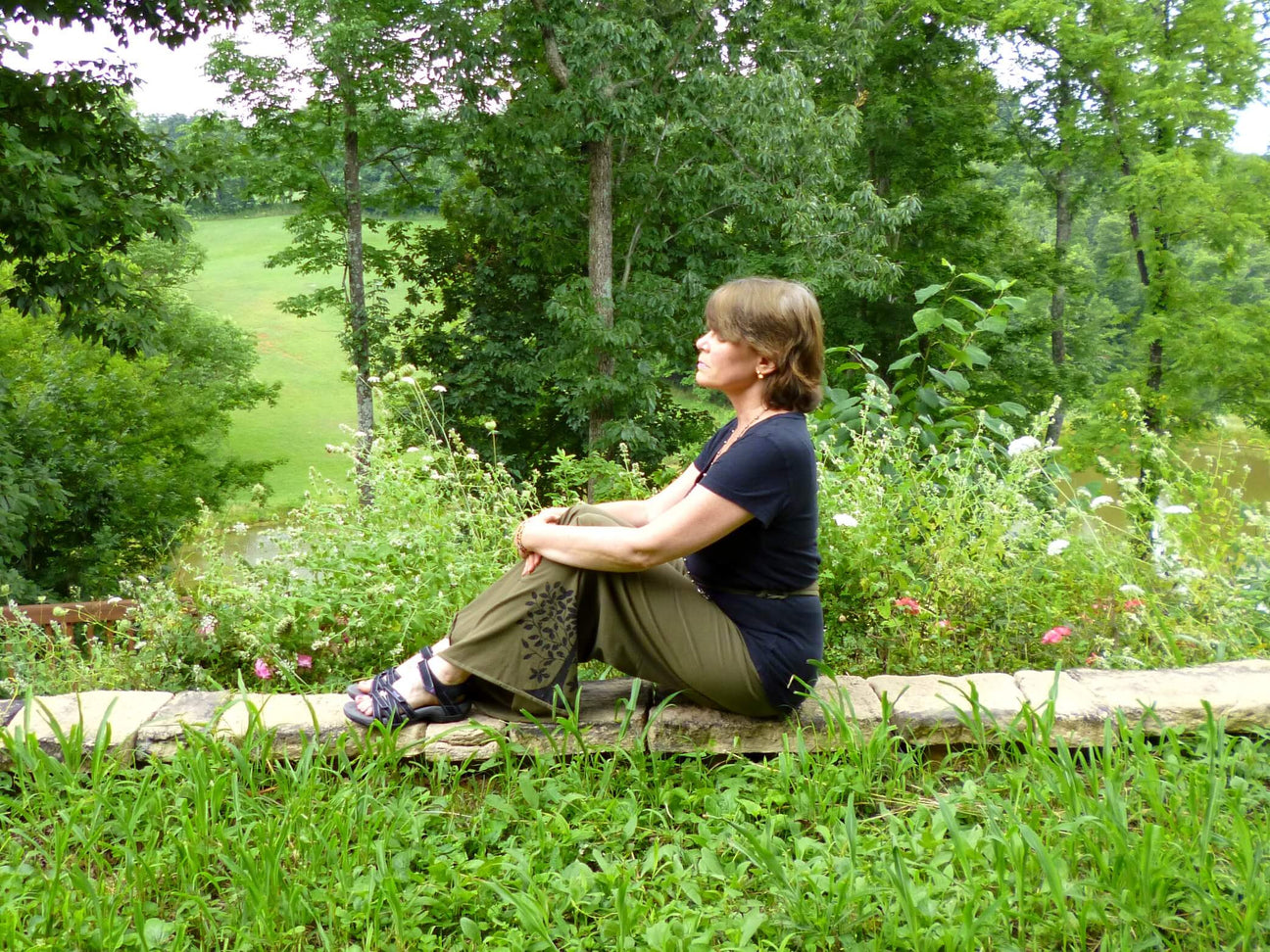 A person enjoys the gift of alone time on a stone ledge in a garden, sitting cross-legged amid lush greenery and wildflowers. Trees and a grassy field stretch peacefully in the background. Soul Flower Blog: Hippie Style & Kind Living