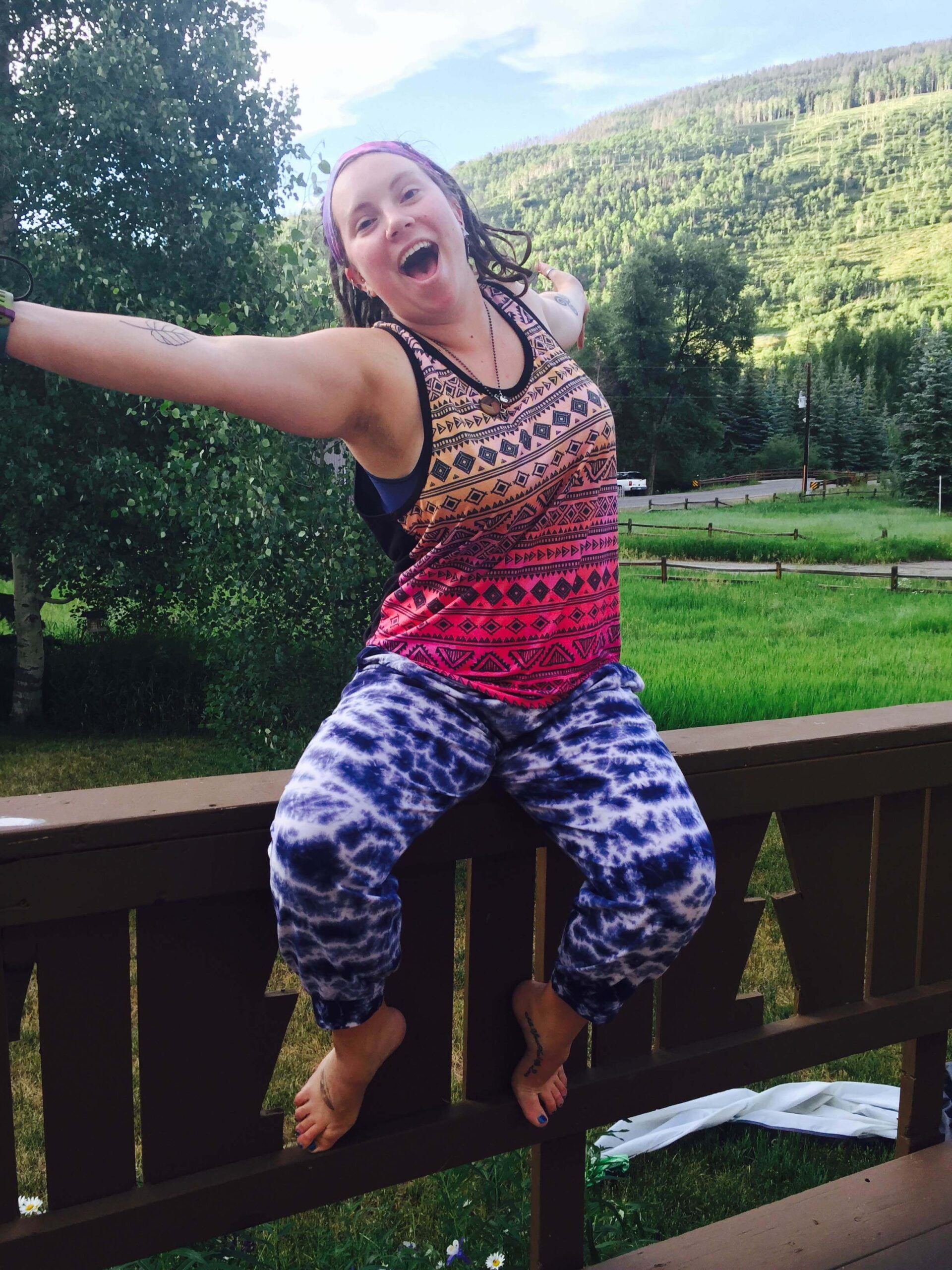 A person with outstretched arms and open mouth sits barefoot on a wooden railing, wearing a colorful tank top and tie-dye pants; green hills and trees are in the background. Soul Flower Blog: Hippie Style & Kind Living