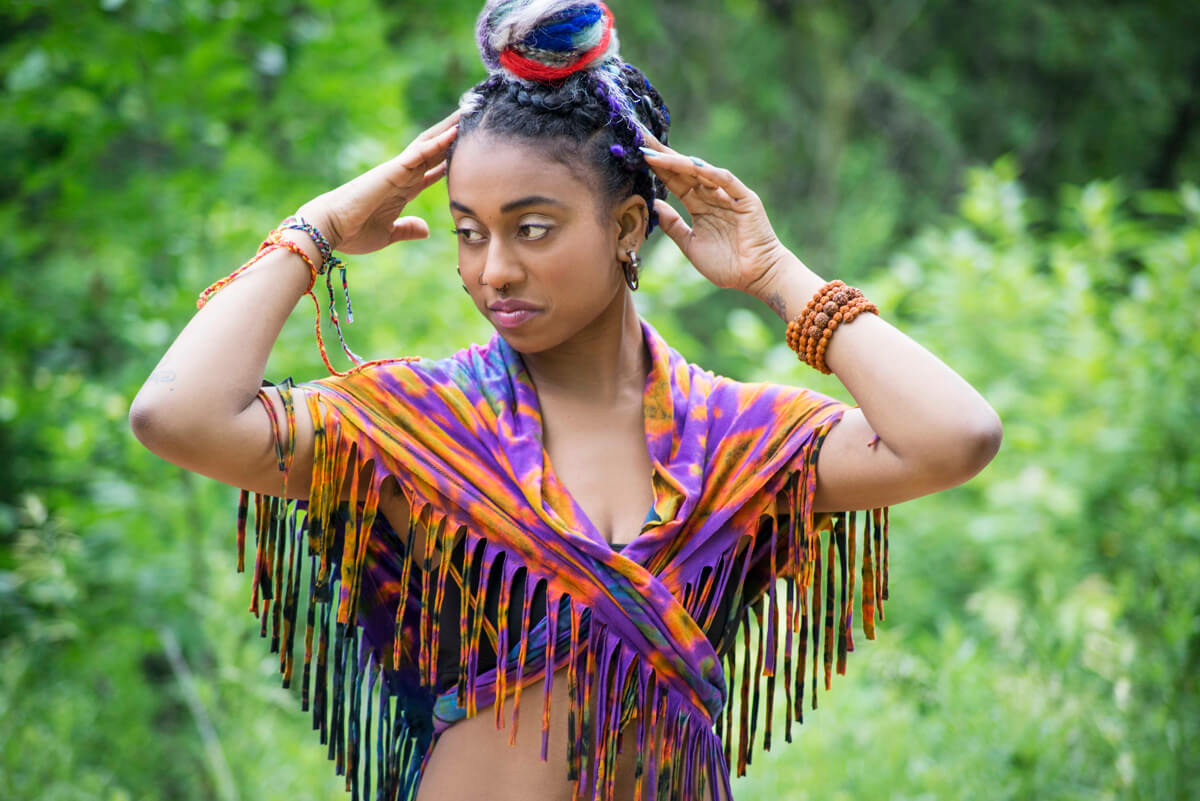 A woman with a colorful, wrapped hairstyle and fringed, multicolored top stands outdoors. Soul Flower Blog: Hippie Style & Kind Living