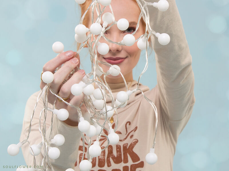 Smiling woman holding a bundle of white string lights in front of a soft blue background, radiating cheerful Hippy Holidays vibes.