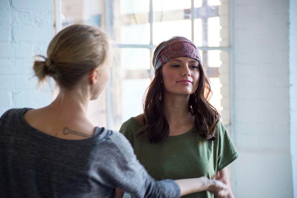 A woman in a green shirt and boho-patterned headband stands indoors while another person gestures toward her. Soul Flower Blog: Hippie Style & Kind Living