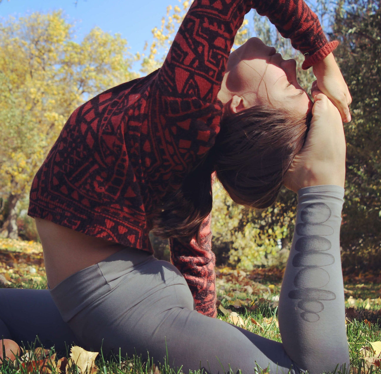 Person practicing a deep yoga stretch outdoors on grass, wearing a funky red sweater and gray leggings, with trees and blue sky in the background for an eco-inspired vibe. Soul Flower Blog: Hippie Style & Kind Living