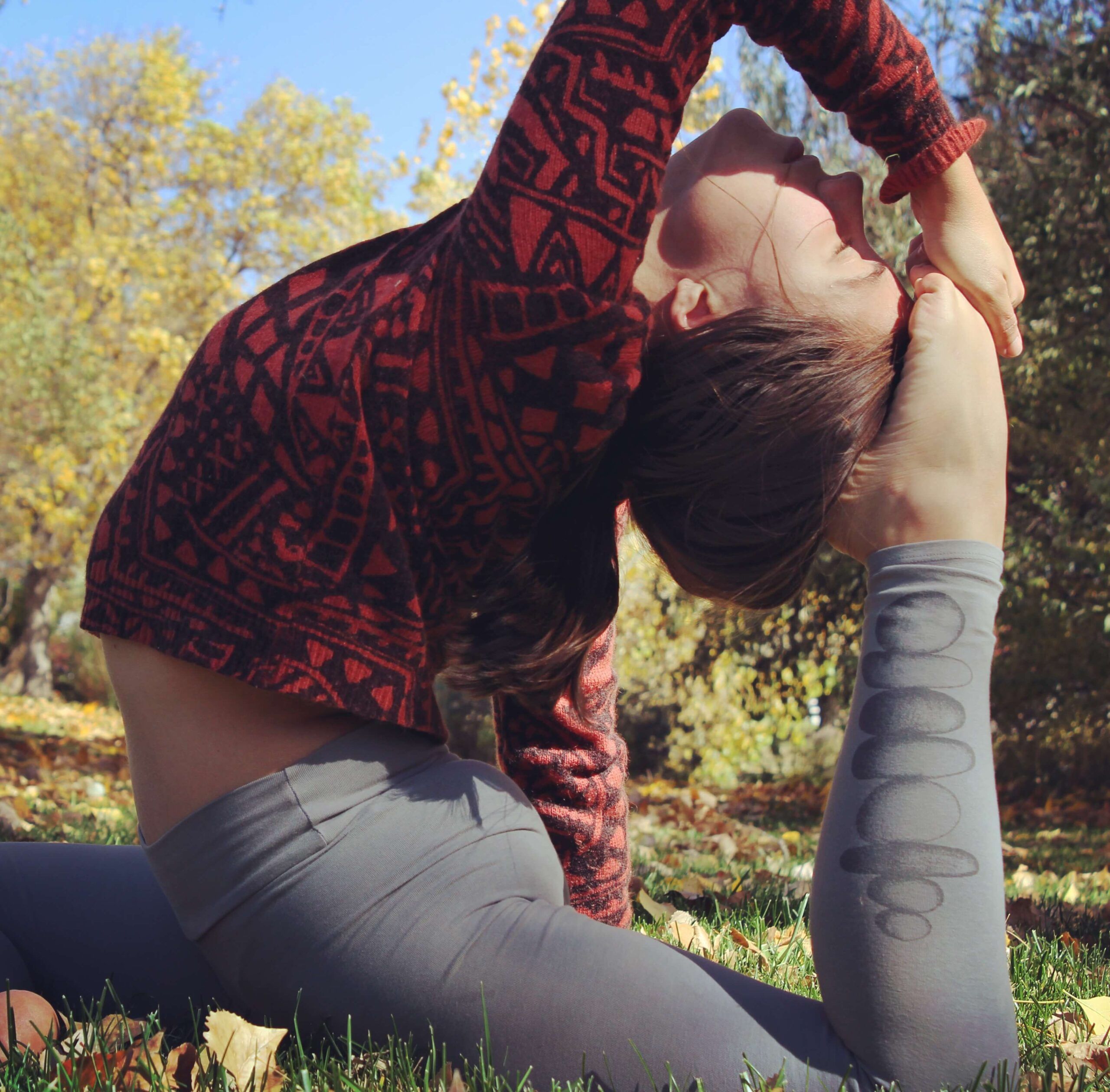 Person practicing a deep yoga stretch outdoors on grass, wearing a funky red sweater and gray leggings, with trees and blue sky in the background for an eco-inspired vibe. Soul Flower Blog: Hippie Style & Kind Living