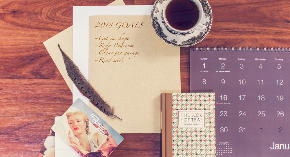 A desk with a Goals Guide for 2018, tea, calendar, books, a feather pen, and vintage postcards.