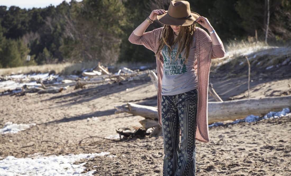 A person with a boho vibe, wearing a hat, long cardigan, patterned pants, and a T-shirt stands on a sandy beach with driftwood and trees in the background. Soul Flower Blog: Hippie Style & Kind Living