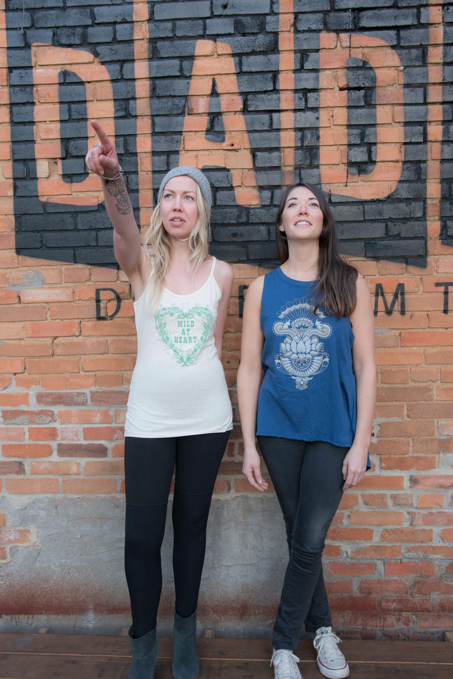Two women stand in front of a brick wall with a painted sign. One woman points forward, while the other stands with her arms at her sides; both gaze ahead with a natural, boho vibe. Soul Flower Blog: Hippie Style & Kind Living