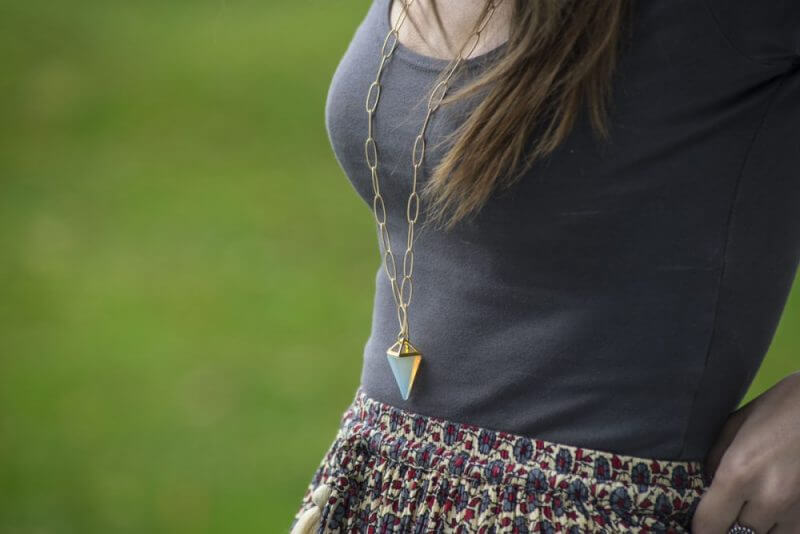 A woman wearing a grey shirt and patterned skirt is shown from the shoulders down, styled with a long chain necklace featuring a geometric pendant for an effortless boho look. Soul Flower Blog: Hippie Style & Kind Living