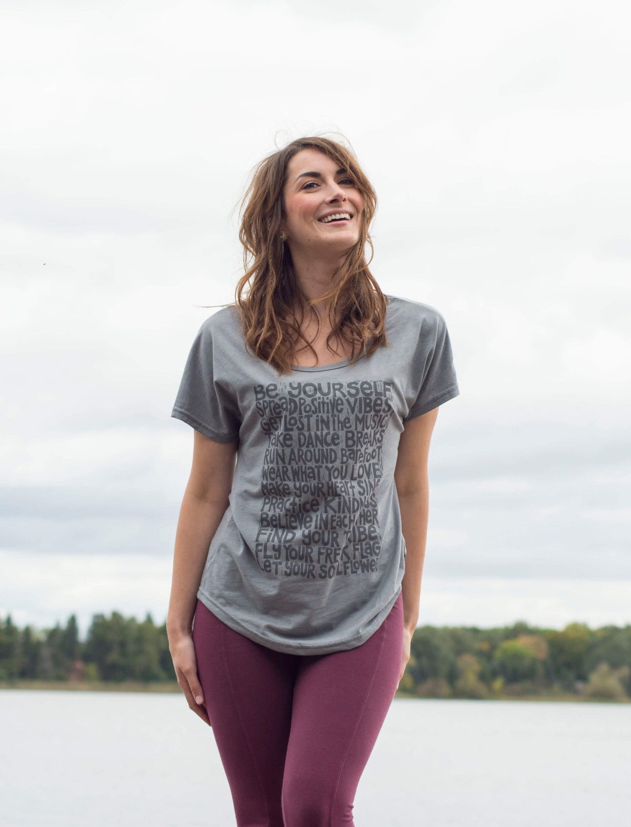 A woman with a boho vibe stands outdoors near a body of water, wearing a gray t-shirt with text and burgundy leggings, smiling with trees and a cloudy sky in the background. Soul Flower Blog: Hippie Style & Kind Living