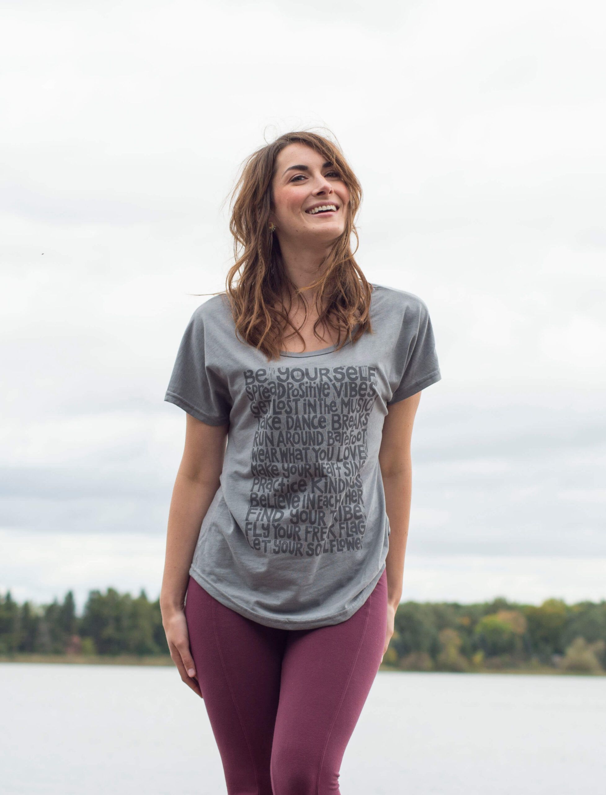 A woman with a boho vibe stands outdoors near a body of water, wearing a gray t-shirt with text and burgundy leggings, smiling with trees and a cloudy sky in the background. Soul Flower Blog: Hippie Style & Kind Living