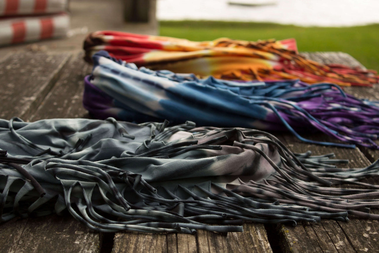 Three fringed, tie-dye scarves in gray, blue, purple, orange, and red are laid out on a weathered wooden table outdoors near a grassy area, adding a vibrant boho touch to your eco-inspired style. Soul Flower Blog: Hippie Style & Kind Living