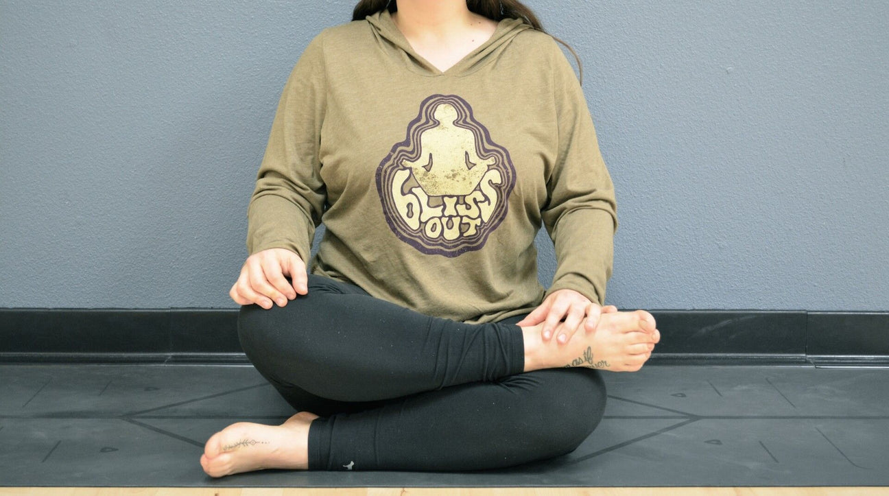 Person sitting cross-legged on a yoga mat against a gray wall, wearing a tan hoodie with a bliss out graphic and black leggings for a cozy, eco-inspired boho vibe. Soul Flower Blog: Hippie Style & Kind Living