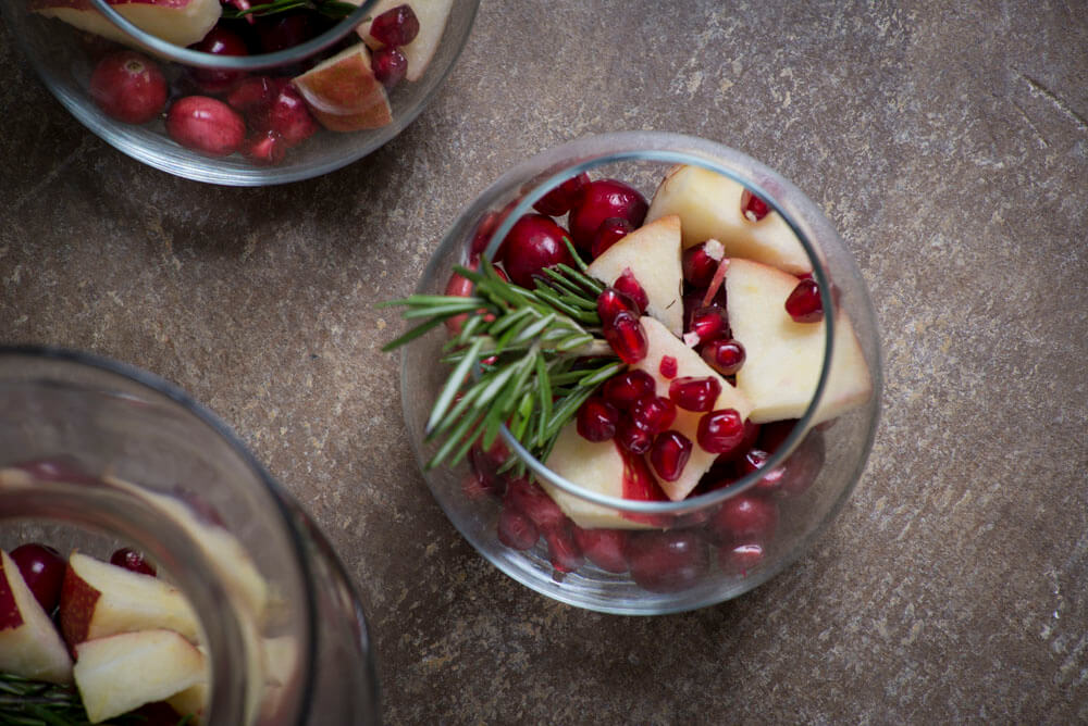 A glass filled with apple slices, pomegranate seeds, cranberries, and a sprig of rosemary sits on a brown surface, adding a touch of boho charm to the earthy scene. Soul Flower Blog: Hippie Style & Kind Living