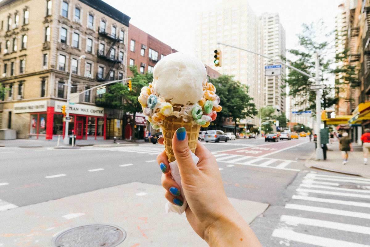 A hand with blue nail polish, wearing a funky hippie t-shirt, holds an ice cream cone topped with colorful cereal on a city street corner with buildings and traffic lights in the background. Soul Flower Blog: Hippie Style & Kind Living