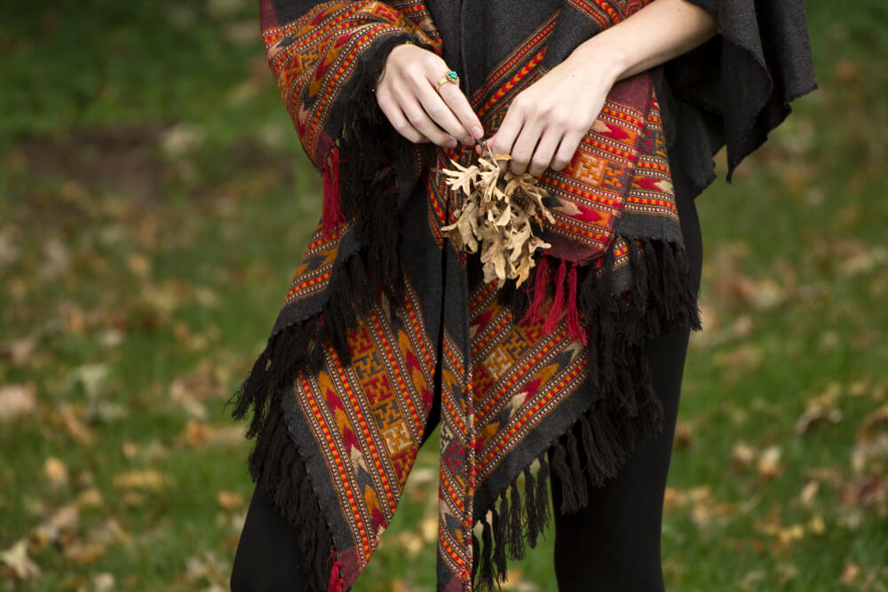 Person wearing an earthy, patterned, fringed shawl stands outdoors on grass, holding a small bunch of dried brown leaves. Soul Flower Blog: Hippie Style & Kind Living