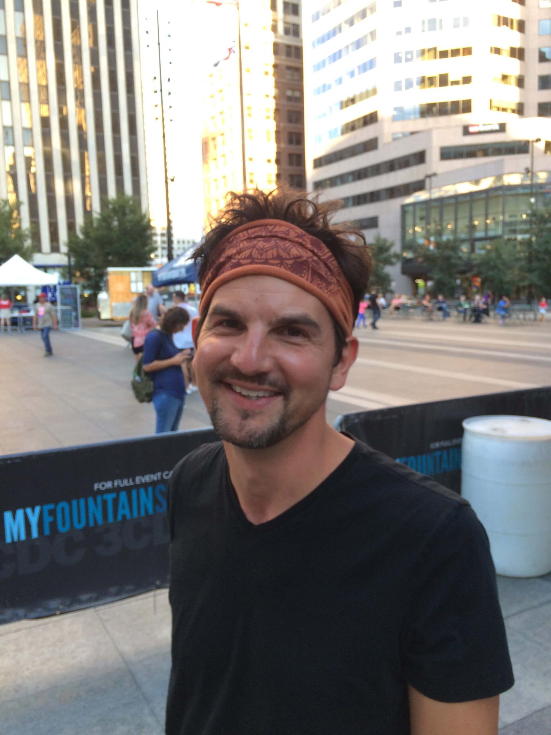 A man wearing a patterned headband and black t-shirt stands outdoors in an urban plaza with tall buildings in the background, embodying a touch of hippie style amid city life. Soul Flower Blog: Hippie Style & Kind Living