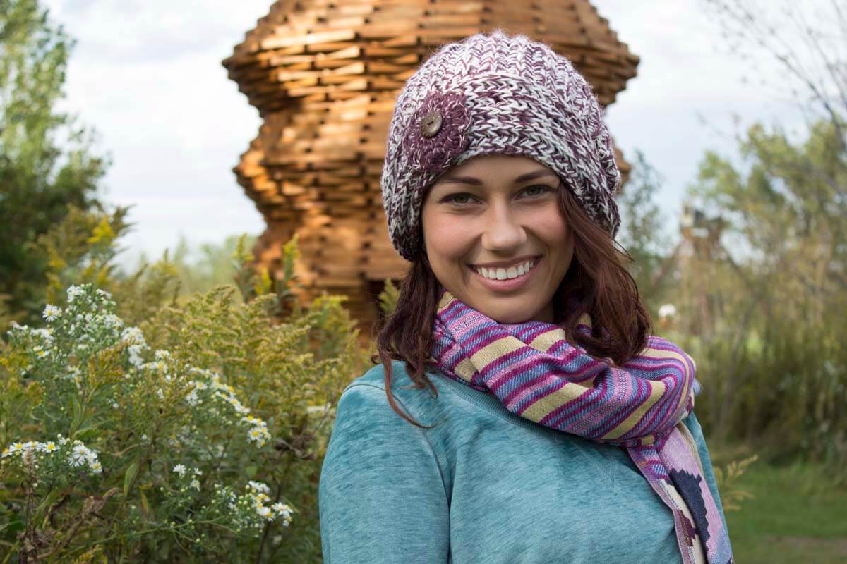 A woman wearing a knit hat and scarf smiles outdoors, standing in front of a wooden sculpture and greenery—captured during our Behind the Scenes: Franconia Photo Shoot Video. Soul Flower Blog: Hippie Style & Kind Living