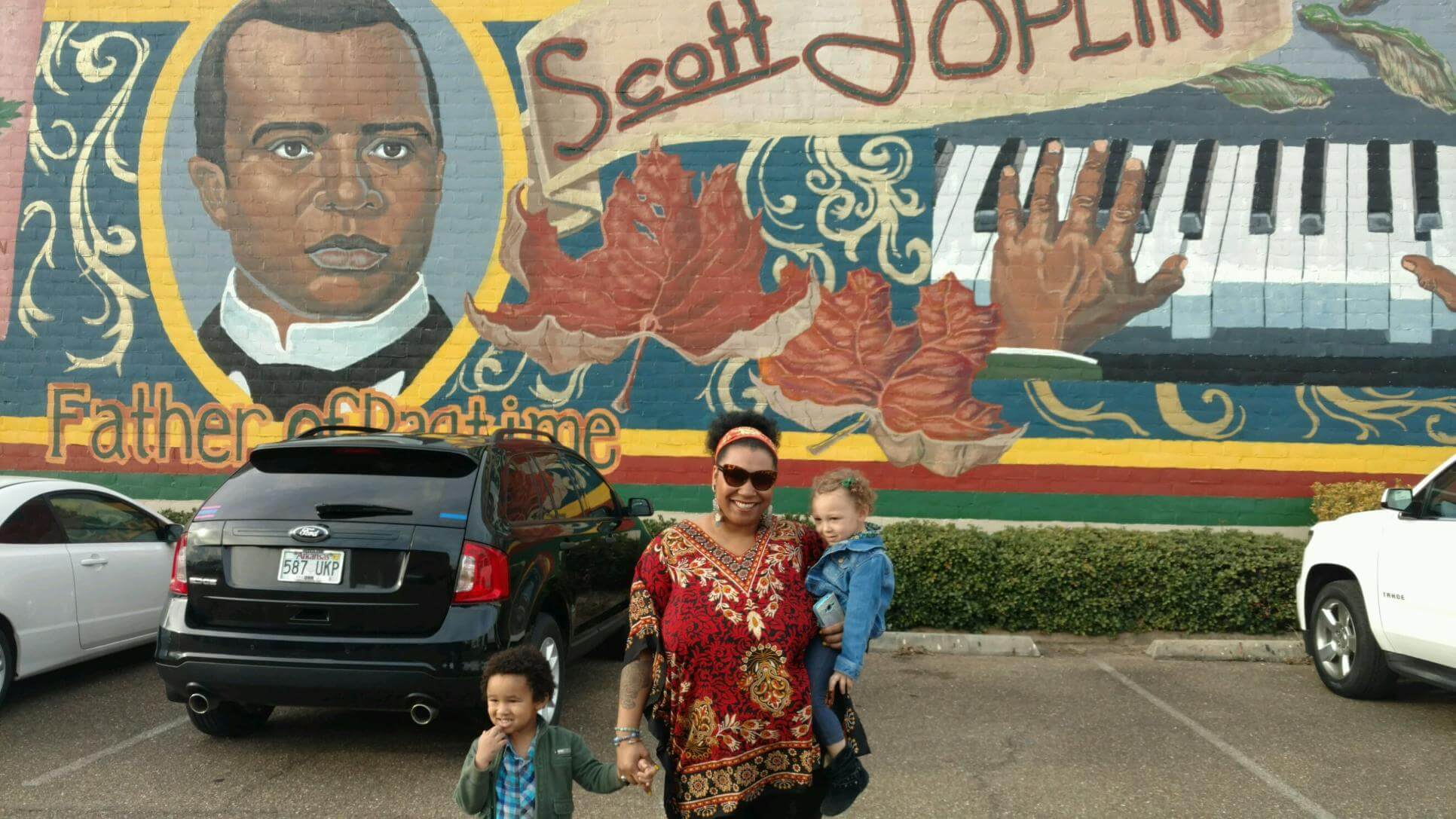A woman and two children stand smiling in front of a boho-style mural of Scott Joplin with a piano and the words “Father of Ragtime.” Cars are parked in front of the mural. Soul Flower Blog: Hippie Style & Kind Living