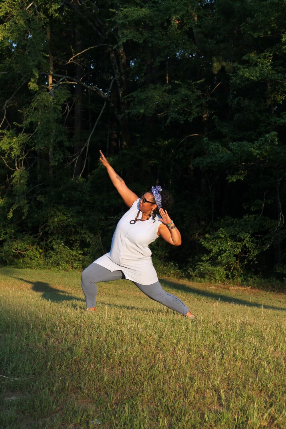A person in a white top and gray leggings strikes a yoga pose on grass, embodying one of the 5 Ways to Become a Bold and Confident Person, with trees behind them and sunlight casting gentle shadows. Soul Flower Blog: Hippie Style & Kind Living