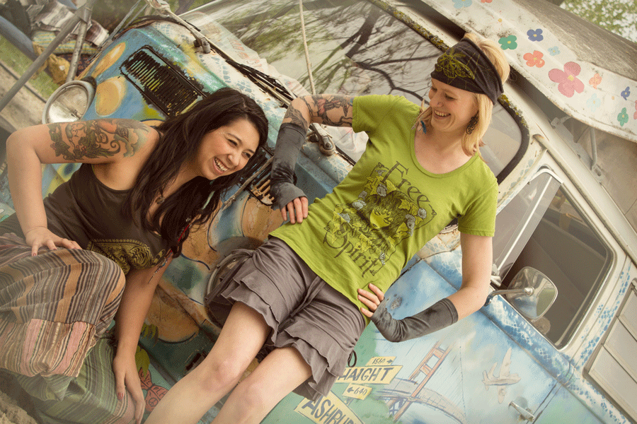 Two women with tattoos smile and lean against a colorful, painted van outdoors. One wears a green Free Spirit shirt; both appear relaxed and happy, radiating an eco-boho vibe perfect for any free-spirited, hippie